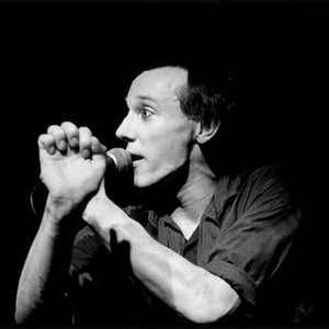 May include: A black and white photo of a person singing into a microphone. The person is wearing a dark shirt and has their hands clasped together.