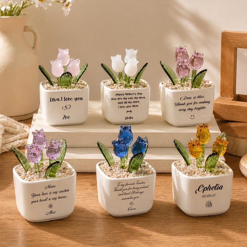 Op de afbeelding: Zes kleine, vierkante witte keramische potten, elk met glazen tulpenbloemen in verschillende kleuren. De potten hebben gepersonaliseerde berichten. De tulpen zijn roze, wit, paars, blauw en geel. De potten staan op een houten oppervlak.
