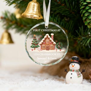 K&ouml;nnte beinhalten: Ein klarer, runder Weihnachtsschmuck mit einem Lebkuchenhaus-Design, einem Weihnachtsbaum und dem Text "FIRST CHRISTMAS AT" und Adressdetails. Der Schmuck h&auml;ngt an einem wei&szlig;en Band. Ein Schneemann befindet sich im Vordergrund.