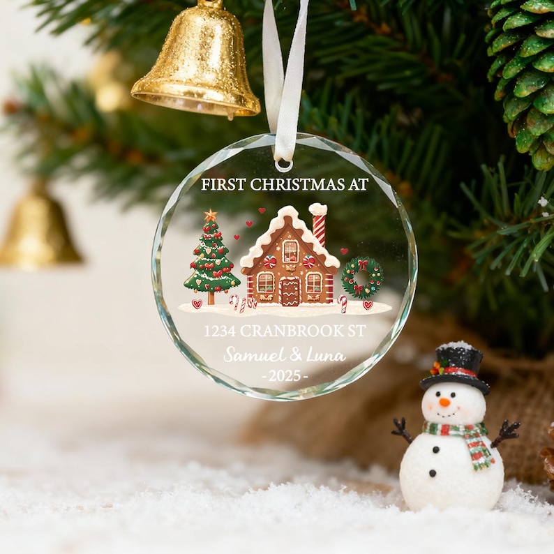 K&ouml;nnte beinhalten: Ein klarer, runder Weihnachtsschmuck mit einem Lebkuchenhaus-Design, einem Weihnachtsbaum und dem Text "FIRST CHRISTMAS AT" und Adressdetails. Der Schmuck h&auml;ngt an einem wei&szlig;en Band. Ein Schneemann befindet sich im Vordergrund.