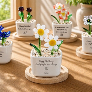 Puede incluir: Maceteros de cerámica blanca con esculturas de flores de cristal. Los maceteros tienen mensajes como "¡Feliz cumpleaños!" y "Agradecido por ti, siempre". Las flores son margaritas, tulipanes y otros diseños coloridos. Cada macetero está sobre un posavasos de madera.