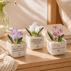 Op de afbeelding: Drie kleine, vierkante witte keramische potten met glazen tulpenbloemen. Elke pot heeft tulpen in een andere kleur: paars, wit en roze. De potten hebben tekst erop, waaronder "Mom", "Stella" en "Lisa".