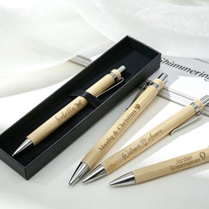 Peut inclure: Ensemble de stylos en bambou personnalisés avec des détails argentés. Un stylo est présenté dans un coffret noir, les autres sont disposés à proximité. Chaque stylo est gravé de noms et d'un petit motif, comme un papillon ou un cœur. Les stylos sont de couleur beige clair.
