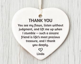 May include: A white heart-shaped ceramic ornament with a cream-colored string for hanging. The text "THANK YOU" is at the top, followed by a heartfelt message and the name "Linda" at the bottom. The ornament is against a light wood background.