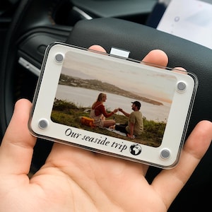 May include: A rectangular photo frame with a white border and clear acrylic front. The frame holds a photo of a couple toasting on a hillside overlooking the ocean. The text "Our seaside trip" is printed at the bottom.