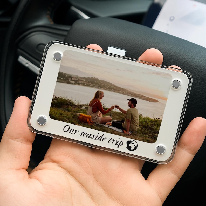 May include: A rectangular photo frame with a white border and clear acrylic front. The frame holds a photo of a couple toasting on a hillside overlooking the ocean. The text "Our seaside trip" is printed at the bottom.