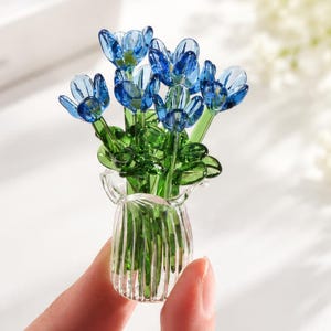 May include: A miniature glass sculpture featuring a bouquet of vibrant blue flowers with green stems and leaves, arranged inside a small, clear, vertically ribbed glass vase. The piece is crafted with translucent glass elements.