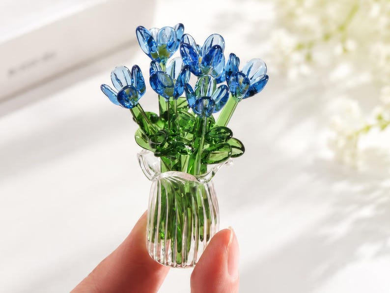 May include: A miniature glass sculpture featuring a bouquet of vibrant blue flowers with green stems and leaves, arranged inside a small, clear, vertically ribbed glass vase. The piece is crafted with translucent glass elements.