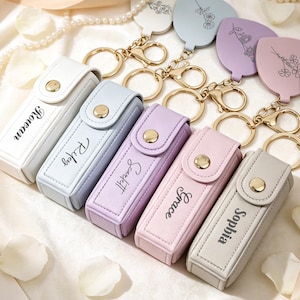 Puede incluir: Una colección de llaveros y estuches de pintalabios personalizados en varios colores pastel. Cada estuche es rectangular con un cierre de botón a presión y un nombre impreso en la parte delantera. También se ven llaveros con forma de flor con herrajes dorados.
