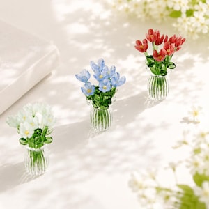 May include: Three miniature glass flower arrangements in clear, ribbed vases. One bouquet features white flowers, one features light blue flowers, and one features red flowers, all with green glass stems and leaves.