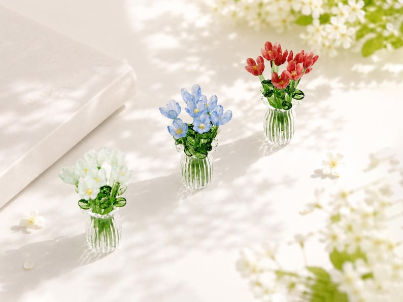 May include: Three miniature glass flower arrangements in clear, ribbed vases. One bouquet features white flowers, one features light blue flowers, and one features red flowers, all with green glass stems and leaves.