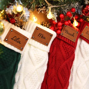 May include: Four Christmas stockings in green, white, and red, with cable knit patterns. Each stocking has a brown leather tag with a name: Lila, Mia, Noah, and Silas. The stockings are hanging on a decorated Christmas tree.