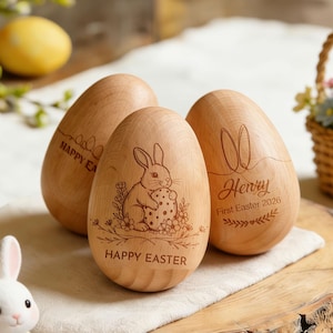 Peut inclure: Trois œufs de Pâques en bois avec des motifs gravés. L'un d'eux représente un lapin tenant un œuf et l'inscription "Happy Easter". Un autre œuf porte des oreilles de lapin et le nom "Henry" avec "First Easter 2026". Un petit panier de fleurs est en arrière-plan.