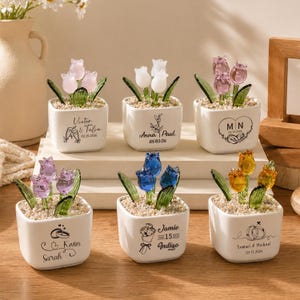 Puede incluir: Seis pequeños maceteros cuadrados de cerámica blanca, cada uno con un arreglo de tulipanes de cristal de diferente color. Las macetas tienen texto decorativo y fechas. Los tulipanes son rosas, blancos, morados, azules y amarillos. Sobre una superficie de madera.