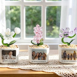 Può includere: Tre sculture di fiori in vetro in vasi quadrati bianchi. Ogni vaso presenta una foto e il testo: "grateful to grow up with you by my side" e "my lifelong best friend". I fiori sono bianchi, rosa e viola.