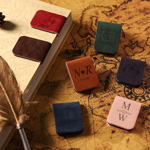 Può includere: Una collezione di segnalibri magnetici in pelle in vari colori, tra cui rosso, marrone, verde, beige, blu navy e rosa. Ogni segnalibro presenta iniziali o date personalizzate. Sono presenti anche una penna e una pigna.