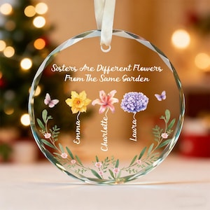 Puede incluir: Adorno redondo de cristal transparente con una cinta blanca para colgar. El adorno presenta el texto "Sisters Are Different Flowers From The Same Garden" e ilustraciones de flores y mariposas. Los nombres "Emma", "Charlotte" y "Laura" están escritos debajo.