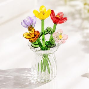 May include: A clear glass vase containing five small, colourful glass flowers. The flowers feature petals in yellow, red, purple, orange, and light pink, each attached to a green glass stem with small green leaves. The vase has a ribbed texture.