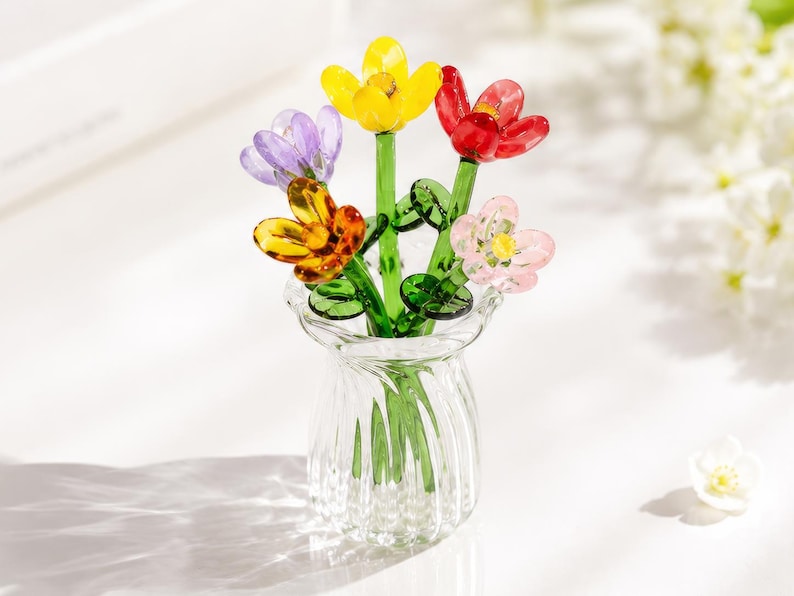 May include: A clear glass vase containing five small, colourful glass flowers. The flowers feature petals in yellow, red, purple, orange, and light pink, each attached to a green glass stem with small green leaves. The vase has a ribbed texture.