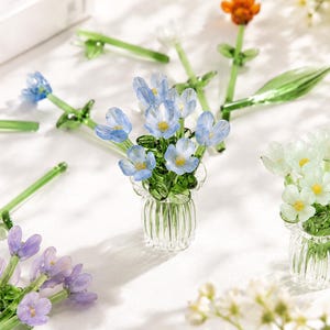 May include: A collection of miniature glass flower arrangements featuring blue, white, and purple blossoms with green stems and leaves, each set in a small, clear, vertically ribbed glass vase, displayed alongside loose glass flowers.