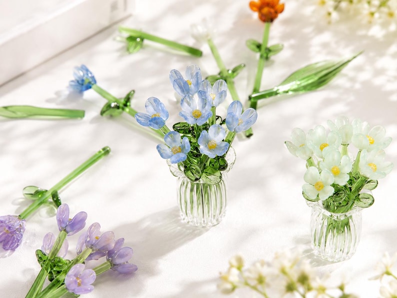 May include: A collection of miniature glass flower arrangements featuring blue, white, and purple blossoms with green stems and leaves, each set in a small, clear, vertically ribbed glass vase, displayed alongside loose glass flowers.