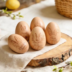 May include: Six wooden Easter eggs with engraved designs, including bunny ears, a bunny, and the words "Easter Happy" and names. The eggs are displayed on a wooden slice and a white cloth. A basket is visible in the background, creating a festive scene.