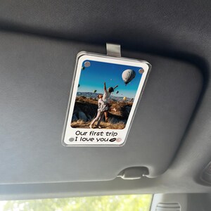 May include: Clear acrylic car visor photo holder with a photo of a couple in front of hot air balloons. The text "Our first trip I love you" is printed at the bottom. A silver clip is attached to the top.