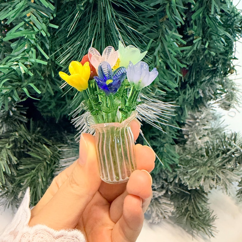 May include: A miniature glass vase containing a bouquet of small, colourful glass flowers in shades of yellow, blue, light purple, pink, and pale green, each featuring green glass stems and leaves, held in a clear, vertically ribbed glass vessel.