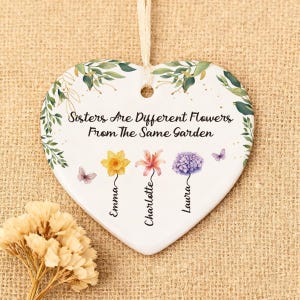 Puede incluir: Adorno de cerámica en forma de corazón con diseño floral y el texto "Sisters Are Different Flowers From The Same Garden". Presenta ilustraciones de flores y los nombres Emma, Charlotte y Laura.