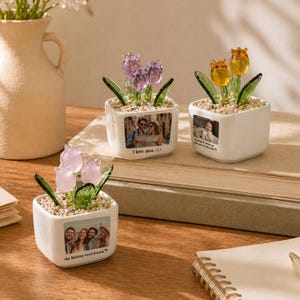 May include: Three square white ceramic planters containing glass flowers and small pebbles. Each planter features a custom photograph on the front. One planter has purple flowers and the text I love you, another has yellow flowers, and one has light pink flowers.