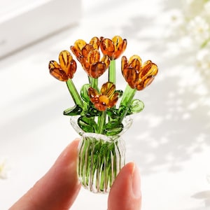 May include: A miniature glass sculpture featuring a cluster of amber-coloured flowers with green stems and leaves, arranged inside a small, clear, vertically ribbed glass vase. The entire piece is crafted from translucent glass materials.