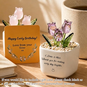 Op de afbeelding: Een decoratief glazen tulpenarrangement in een witte vierkante pot met kleine steentjes. Het arrangement bevat drie paarse tulpen en groene bladeren. Een kaart leest "Happy Early Birthday! Love from your Sister".