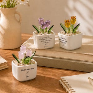 Op de afbeelding: Drie kleine, witte vierkante keramische potten met glazen tulpenbloemen in roze, paarse en gele tinten. De potten hebben tekst op de voorkant, waaronder "Lisa, I love you" en "To my favorite teacher." De tulpen staan in een bedje van kleine steentjes.