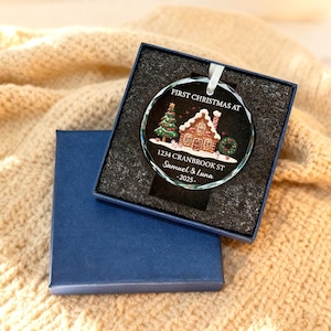 K&ouml;nnte beinhalten: Ein klarer Weihnachtsbaumschmuck aus Glas mit einem Lebkuchenhaus-Design, einem Weihnachtsbaum und dem Text "FIRST CHRISTMAS AT" sowie personalisierten Details. Der Schmuck befindet sich in einer blauen Geschenkbox.