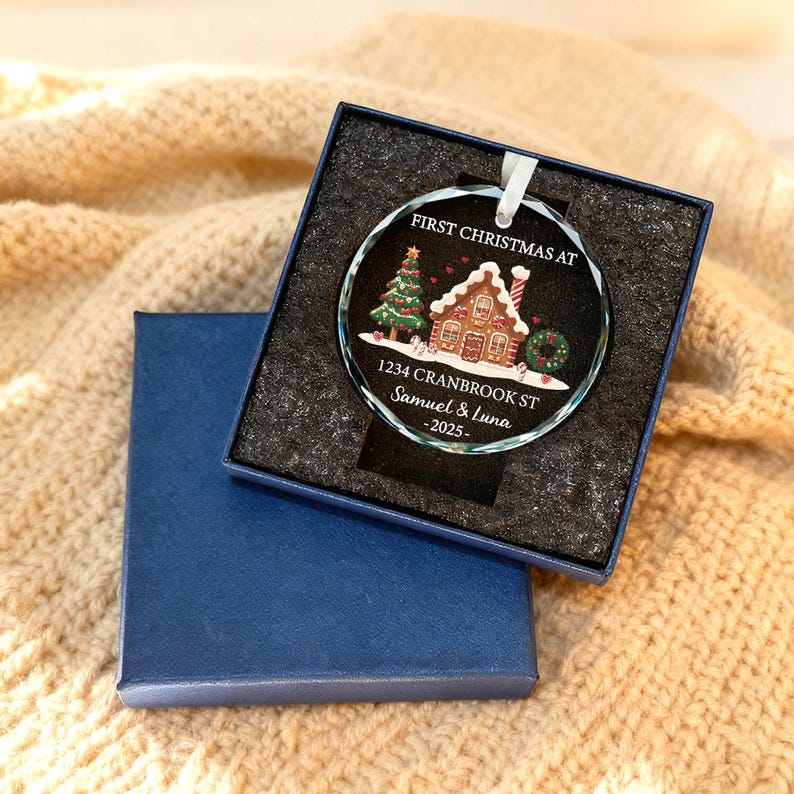 K&ouml;nnte beinhalten: Ein klarer Weihnachtsbaumschmuck aus Glas mit einem Lebkuchenhaus-Design, einem Weihnachtsbaum und dem Text "FIRST CHRISTMAS AT" sowie personalisierten Details. Der Schmuck befindet sich in einer blauen Geschenkbox.
