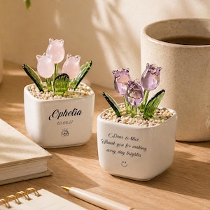 Op de afbeelding: Twee kleine, witte vierkante keramische potten met glazen tulpenbloemen. E&eacute;n pot heeft roze tulpen en de naam "Ophelia" met een datum en een taartpictogram. De andere pot heeft paarse tulpen en de tekst "Dear Alice. Thank you for making every day brighter".