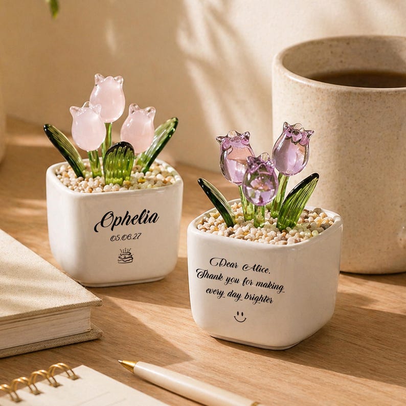 Op de afbeelding: Twee kleine, witte vierkante keramische potten met glazen tulpenbloemen. E&eacute;n pot heeft roze tulpen en de naam "Ophelia" met een datum en een taartpictogram. De andere pot heeft paarse tulpen en de tekst "Dear Alice. Thank you for making every day brighter".