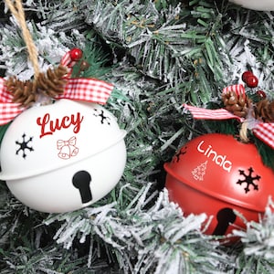 May include: Two Christmas bell decorations, one white and one red, hang on a decorated tree. The white bell has "Lucy" in red script, and the red bell has "Linda". Both have snowflake cutouts and are adorned with red and white checked bows.