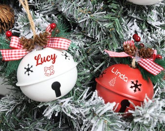 May include: Two Christmas bell ornaments, one white and one red, hang on a decorated tree. The white bell has "Lucy" in red script, and the red bell has "Linda". Both have snowflake cutouts and are adorned with red and white checkered bows.