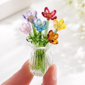 May include: A miniature glass vase containing a bouquet of small, multi-coloured glass flowers. The flowers feature petals in shades of purple, light pink, blue, red, yellow, and orange, attached to green glass stems with small green leaves.