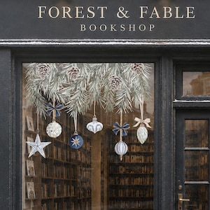 Peut inclure: La devanture de la librairie Forest & Fable présente une façade noire avec des lettres dorées. La grande vitrine est décorée de branches de pin suspendues et de divers ornements, dont une étoile blanche et des boules bleues et blanches.
