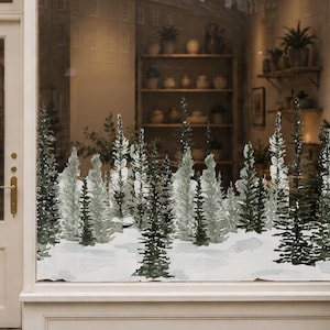 Könnte beinhalten: Ein Ladenlokal mit cremefarbener Fassade und einem Schild mit der Aufschrift Willow & Wild HOMEWARES. Das große Schaufenster zeigt einen Winterwald-Aufkleber mit verschneiten Bäumen. Zwei Topf-Tannen flankieren den Eingang und eine Girlande hängt darüber.