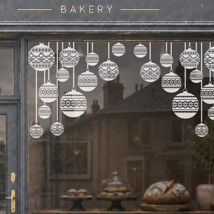 May include: The storefront of The Flour Pot bakery features a dark grey facade with a large window decorated with white hanging ornament decals. A wooden A-frame sign on the pavement lists SOURDOUGH, PASTRIES, COFFEE, and CAKE in white text.
