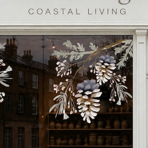 Puede incluir: Un adhesivo para ventana que presenta dos ramas simétricas con agujas de pino, pequeñas bayas y grandes piñas en tonos blanco, gris y azul. El diseño está aplicado en el cristal de un escaparate con el texto COASTAL LIVING encima.