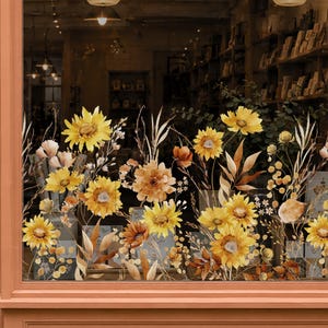 May include: A window decal featuring a vibrant arrangement of yellow sunflowers, golden wildflowers, and dried grasses. The floral design displays various shades of yellow, orange, and brown, creating a dense, botanical display across the glass surface.