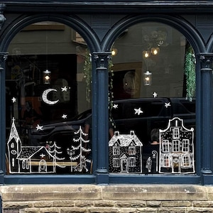 Könnte beinhalten: Ein Schaufenster mit weißen Strichzeichnungen einer Kirche, Häusern, Bäumen, einem Halbmond und Sternen. Das Fenster wird von dunkelblauen Bögen und Säulen mit einem Steinsockel umrahmt.