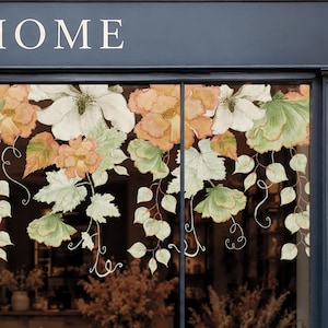 Könnte beinhalten: Ein Fensteraufkleber mit einem Blumenarrangement im Aquarellstil, bestehend aus großen weißen Blüten, zart orangefarbenen Blumen und hellgrünen Blättern mit feinen Ranken, angebracht auf einer Fensterscheibe mit einem dunklen Rahmen.