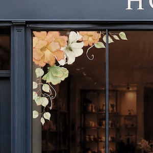 May include: A floral window decal featuring watercolour-style orange and white blossoms with green leaves and curling vines. The design is arranged in a corner cluster, displaying soft colour gradients and organic shapes applied to a glass surface.