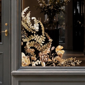 Könnte beinhalten: Ein dekorativer Fensteraufkleber mit einem botanischen Arrangement aus goldfarbenen Farnen, Tannenzapfen und kleinen Blüten. Das Klebedesign ist in der Ecke einer Glasscheibe angebracht und zeigt detaillierte Blattmuster und Strukturen.