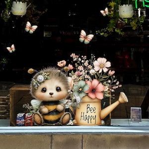 Op de afbeelding: Een grillige illustratie toont een cartoonbij met een bloemenkroon, zittend naast een gieter gevuld met bloemen. Op de gieter staat "Bee Happy". Vlinders en hangplanten dragen bij aan de vrolijke scène.